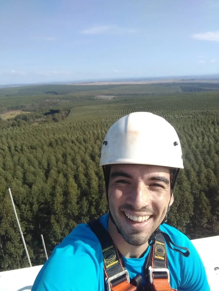 Selfie on top of a camera tower, wearing a helmet and harness, with a eucalyptus plantation stretching to the horizon.